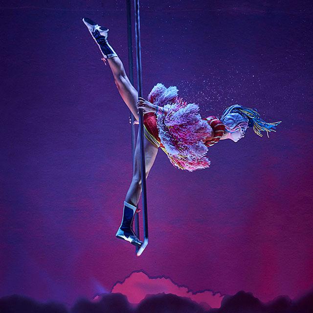 Un artiste du Cirque du Soleil spécialisé dans les figures aériennes à la barre fixe tourne et se balance la tête en bas dans un costume coloré sous un éclairage violet.