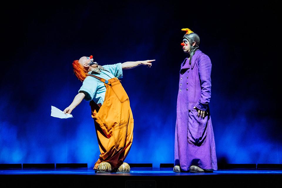 Deux clowns du Cirque du Soleil interprètent un sketch ludique sur scène, vêtus de costumes colorés sous un éclairage théâtral bleu.