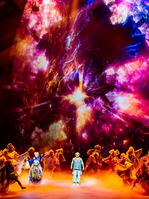 La troupe du Cirque du Soleil se produit devant un décor numérique spectaculaire de style cosmique, avec des couleurs vives et de la fumée sur scène.
