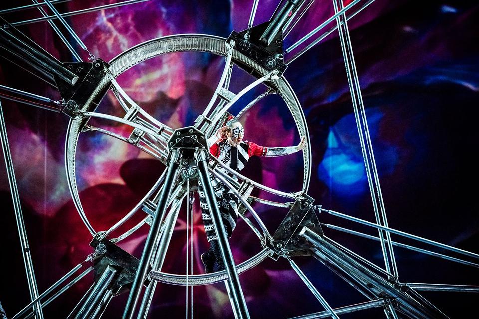 La troupe du Cirque du Soleil se produit devant un décor numérique spectaculaire de style cosmique, avec des couleurs vives et de la fumée sur scène.