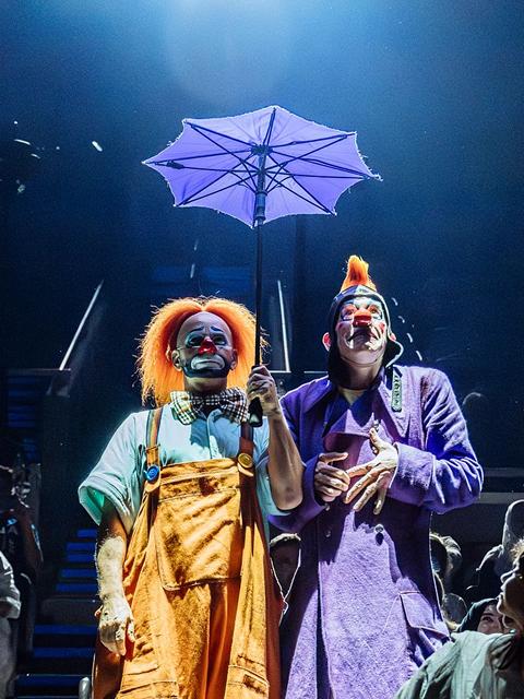 Deux clowns du Cirque du Soleil se tiennent sur scène, tenant un parapluie, vêtus de costumes colorés sous un éclairage théâtral bleu doux.