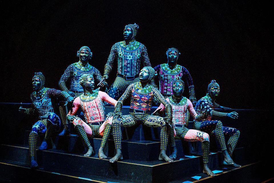 Les artistes du Cirque du Soleil, vêtus de costumes détaillés posent ensemble sur des plateformes à plusieurs niveaux sous un éclairage spectaculaire.