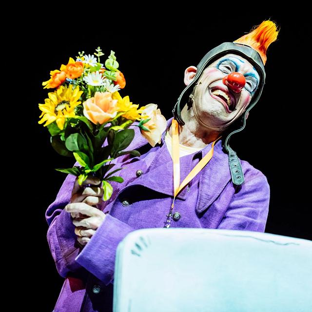 Un clown du Cirque du Soleil vêtu d'un costume violet sourit tout en tenant un bouquet de fleurs jaunes et orange sur scène.