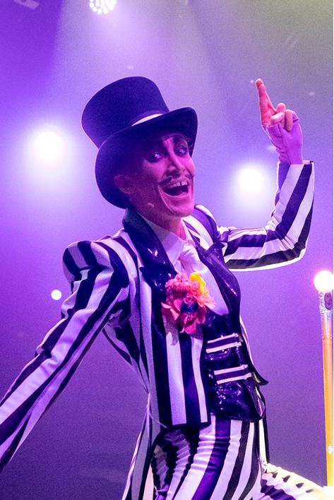 Un artiste du Cirque du Soleil vêtu d'un costume rayé noir et blanc et d'un haut-de-forme sourit et gesticule de manière enjouée sous les lumières colorées de la scène.