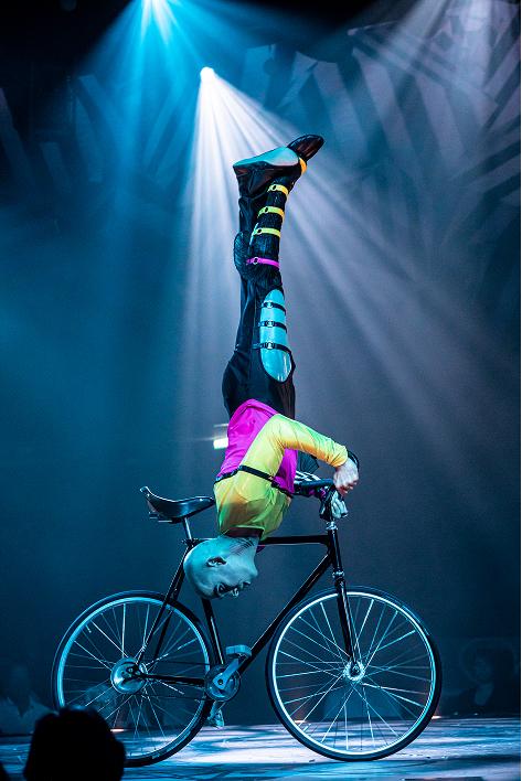 Un acrobate du Cirque du Soleil réalise un équilibre sur les mains sur un vélo sous un éclairage bleu spectaculaire.