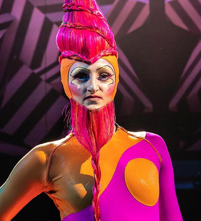 Portrait en gros plan d'un artiste du Cirque du Soleil vêtu d'un costume rose et orange vif, avec un maquillage théâtral audacieux et une coiffure sculpturale.