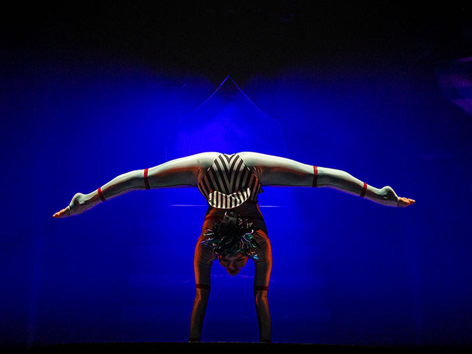 Une contorsionniste du Cirque du Soleil réalise un grand écart arrière sur fond bleu foncé.