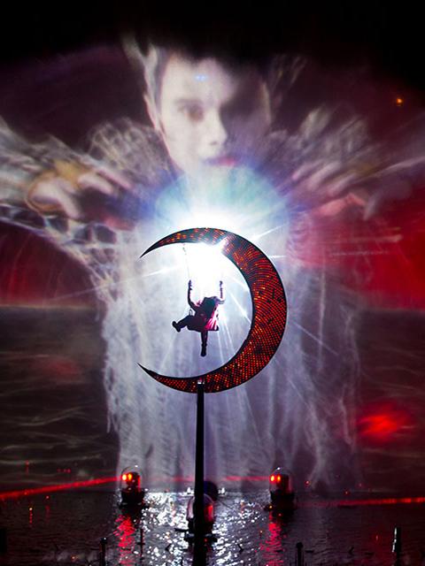 Cirque du Soleil aerial performer suspended on a glowing crescent moon in front of dramatic projected visuals and red lighting.