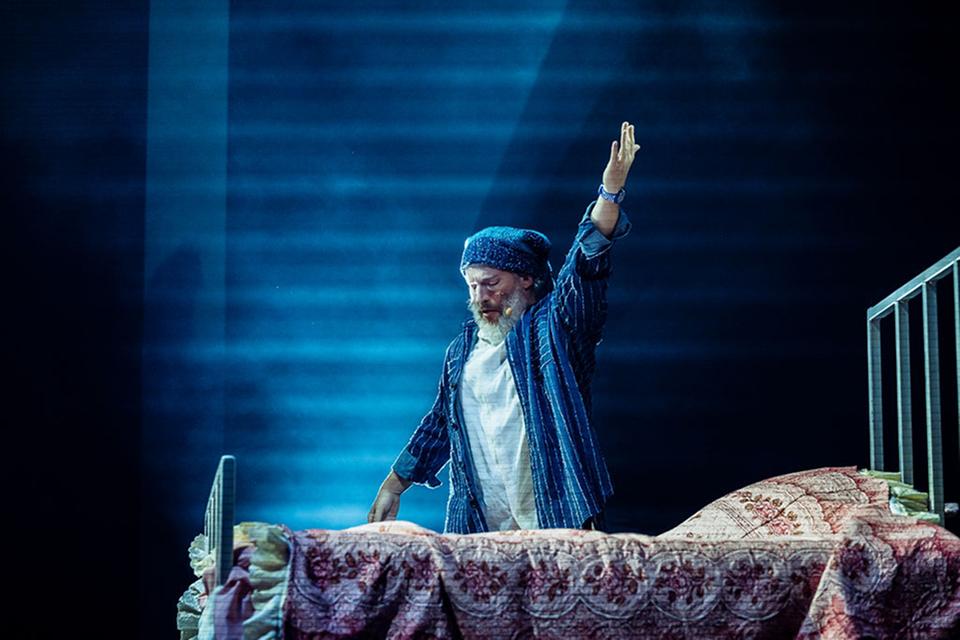 Un artiste du Cirque du Soleil vêtu d'un pyjama rayé lève la main alors qu'il se tient debout sur un lit sous un éclairage bleu.