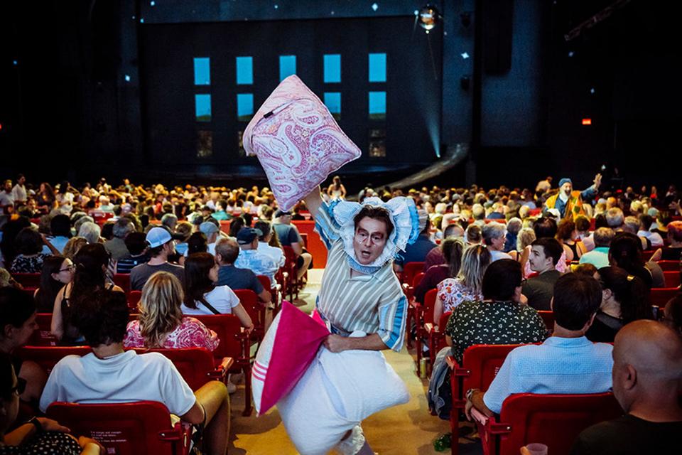 Un personnage du Cirque du Soleil transporte des coussins géants parmi les spectateurs assis, interagissant de manière ludique avec eux.