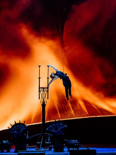 Un acrobate du Cirque du Soleil réalise un saut en hauteur et une figure à la perche devant un décor de scène orange et rouge flamboyant.