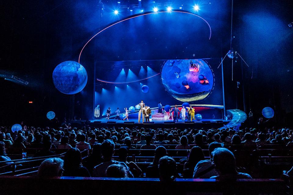 Vue panoramique d'un spectacle du Cirque du Soleil dans une arène, avec des appareils aériens, des sphères flottantes et un public nombreux sous un éclairage bleu.