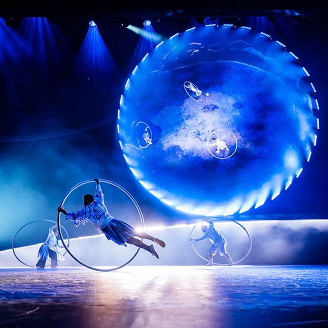 Les acrobates du Cirque du Soleil exécutent des figures sur des roues Cyr devant un décor circulaire lumineux, sur une scène éclairée de bleu.