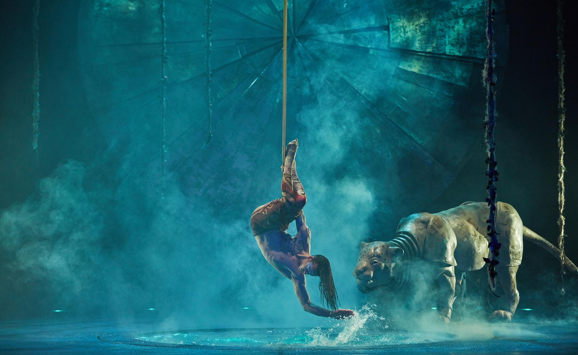 A Cirque du Soleil aerial performer hangs upside down from a rope above water, performing beside a life-sized tiger sculpture in a dramatic, mist-filled stage setting.