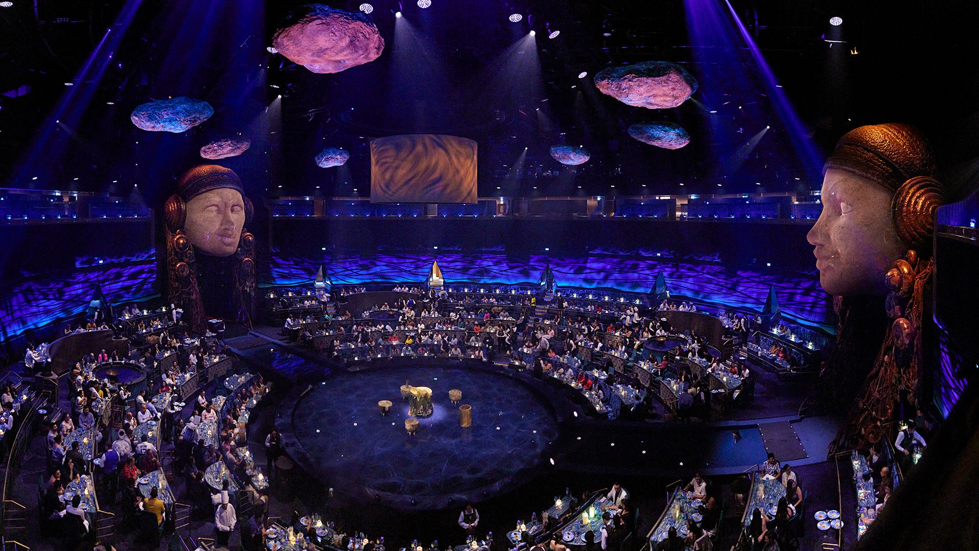 Panoramic view of a large immersive dinner theater with guests seated around a circular stage, dramatic purple lighting, floating rock-like set pieces, and monumental sculpted heads overlooking the audience.