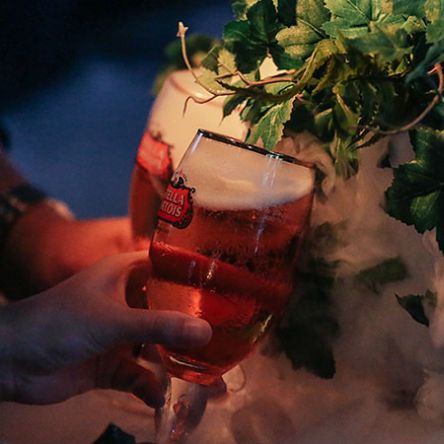 Close-up of a hand holding a glass of amber beer with foam, set in ice beside fresh green herbs in a bar setting.