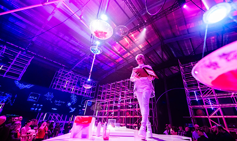 Immersive stage set with blue and purple lighting, featuring large scaffold structures, elevated platforms, suspended mechanical elements, and performers moving across a futuristic theatrical environment.