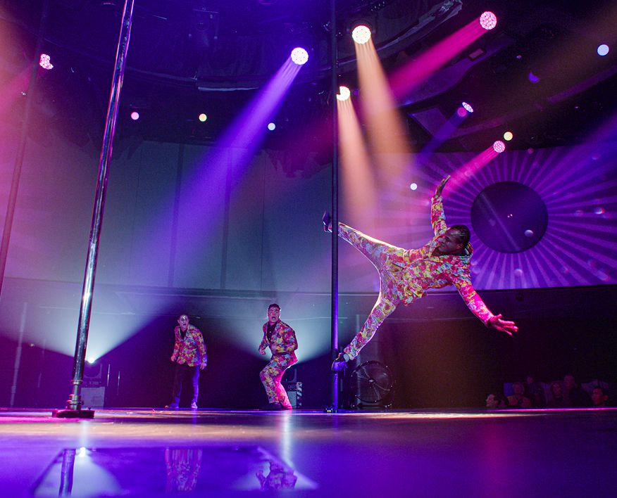 Cirque du Soleil acrobat performs a dynamic aerial move on a pole while other performers dance on a brightly lit stage with purple and gold spotlights.