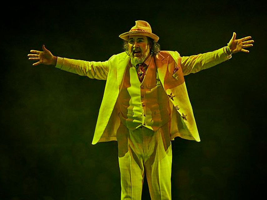 Cirque du Soleil clown character in a bright yellow suit stands center stage with arms open under dramatic spotlight.