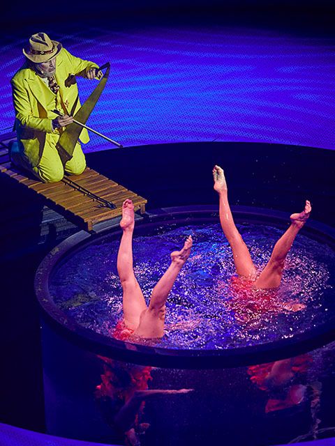 Cirque du Soleil performers execute a synchronized water acrobatics routine in a circular pool while a musician plays above them.