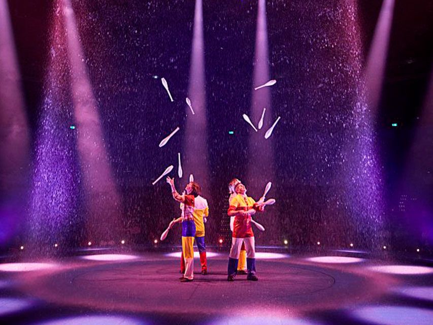 Cirque du Soleil jugglers perform with white clubs under pink and purple stage lights in a theatrical arena.