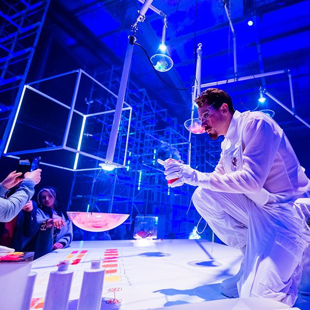 Un artiste du Cirque du Soleil vêtu d'un costume blanc prépare une expérience théâtrale sur une scène interactive entourée d'invités sous un éclairage bleu.