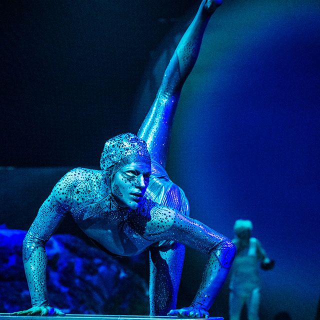 Une contorsionniste du Cirque du Soleil vêtue d'un costume bleu scintillant exécute une pose spectaculaire au sol sous les projecteurs de la scène.