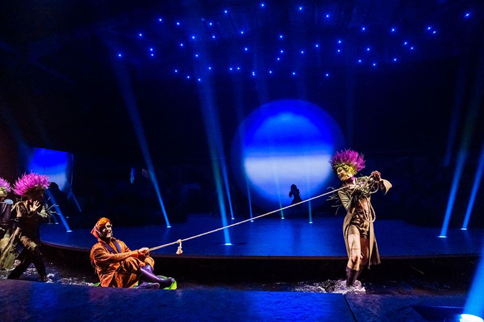 Les artistes du Cirque du Soleil jouent une scène ludique sur une scène aquatique à l'aide de cordes et d'accessoires, sous un éclairage bleu étoilé.