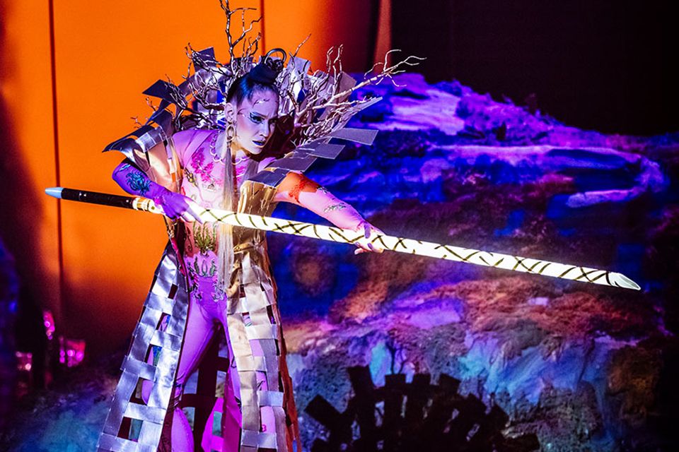 Un artiste du Cirque du Soleil vêtu d'un costume sophistiqué brandit un bâton lumineux sur une scène aux couleurs vives et à la scénographie spectaculaire.