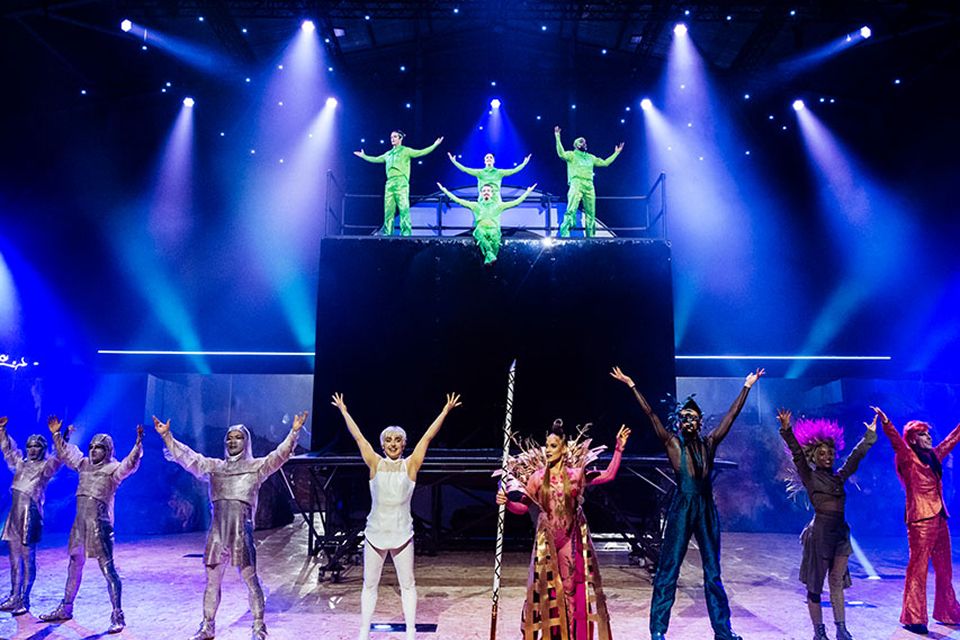 Les artistes du Cirque du Soleil saluent le public sur scène, levant les bras sous les projecteurs bleus et violets.